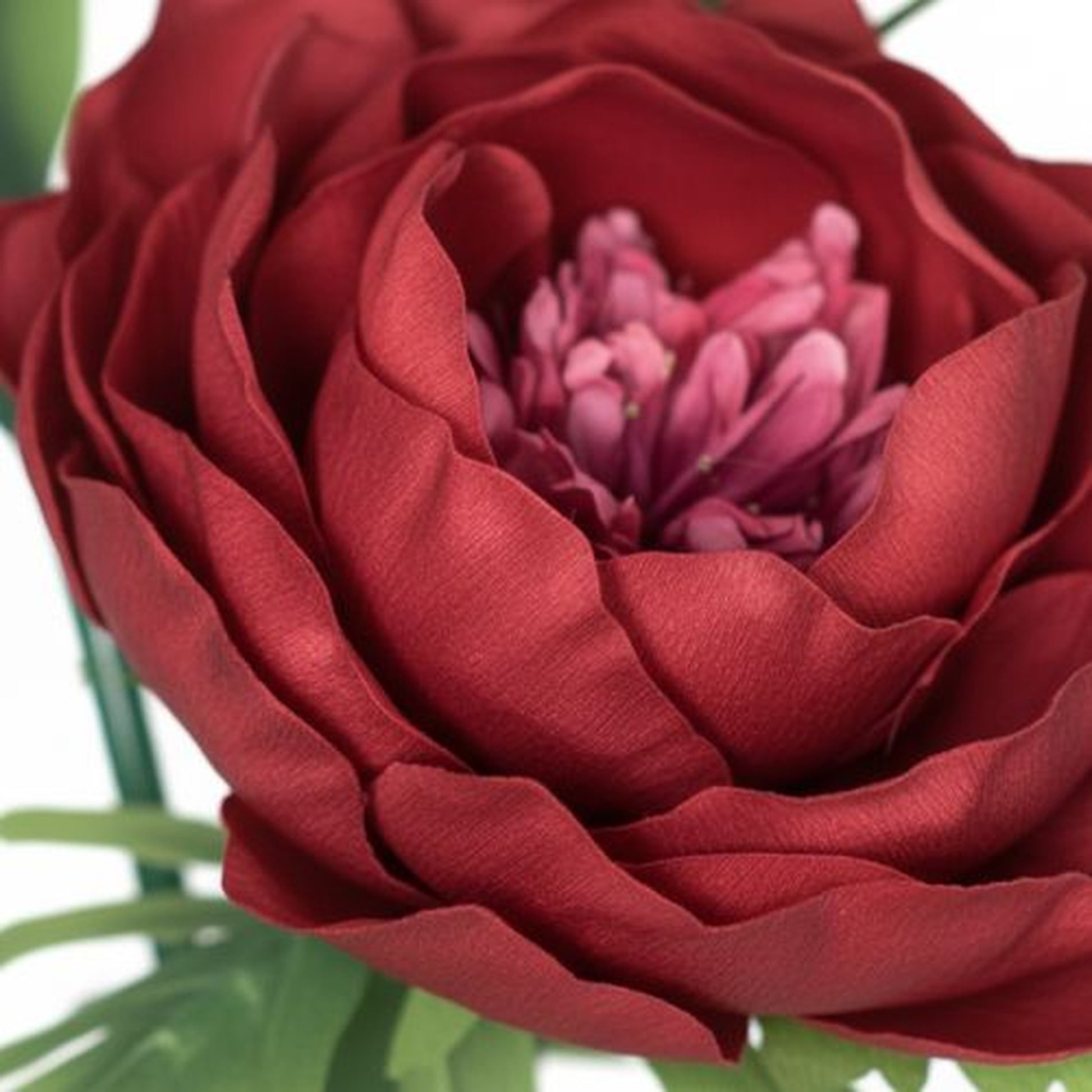 "Extreme macro close-up photograph of a single artificial flower resembling a peony or ranunculus. Deep crimson red outer petals surround a dense cluster of smaller, frilly magenta and pink petals in the center. Fine fabric texture visible on the petals arranged in soft overlapping layers. Shallow depth of field with sharp focus on the flower center and blurred green foliage in background. White backdrop, professional product photography showcasing artisan craftsmanship."