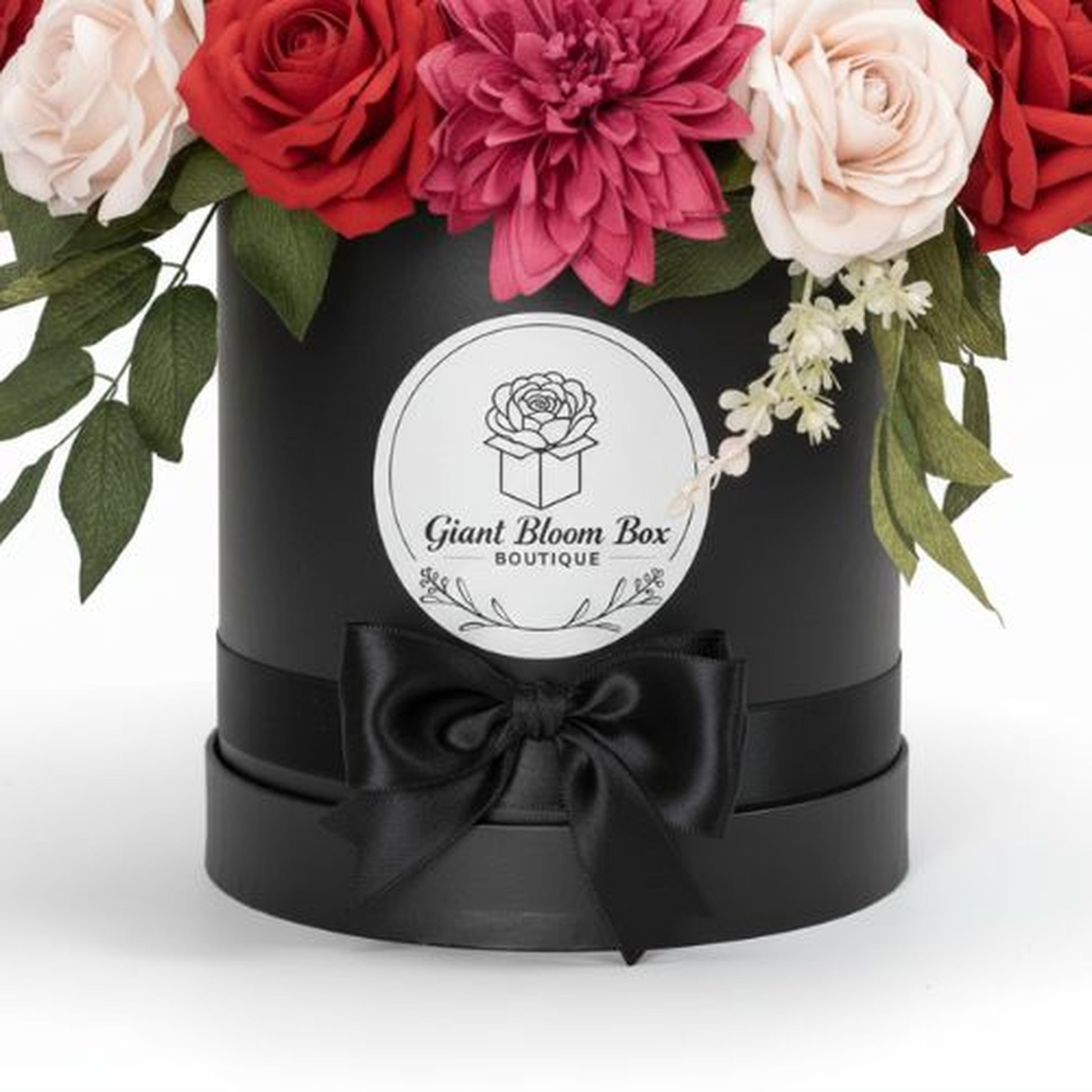 "High-quality studio photograph of a black cylindrical gift box containing an elegant floral arrangement. The matte black box features a circular white label on the front with black line-art logo of a rose emerging from an open box, text reading 'Giant Bloom Box' in script font and 'BOUTIQUE' below. A wide black satin ribbon is wrapped around the box and tied in a bow. The arrangement includes deep red roses, pale pink roses, a large magenta dahlia-like flower, dark green leaves, and delicate white blossoms