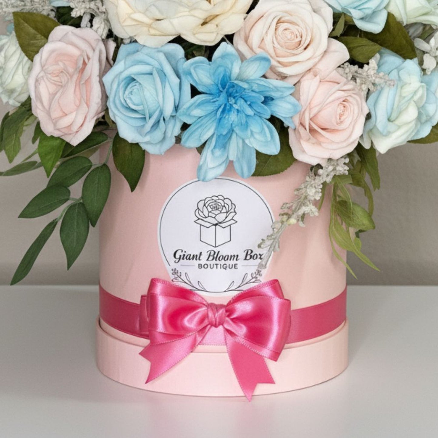 Professional product photograph of an elegant dome-shaped floral arrangement in a light pink cylindrical bloom box. Dense bouquet features artificial roses in soft pastel blue, blush pink, and creamy ivory, along with vibrant sky blue dahlia or chrysanthemum. Dark green eucalyptus leaves on left side, delicate frosted baby's breath filler flowers throughout. The box has wide lustrous hot pink satin ribbon wrapped around it tied in large multi-looped bow at front. White circular label on front displays black