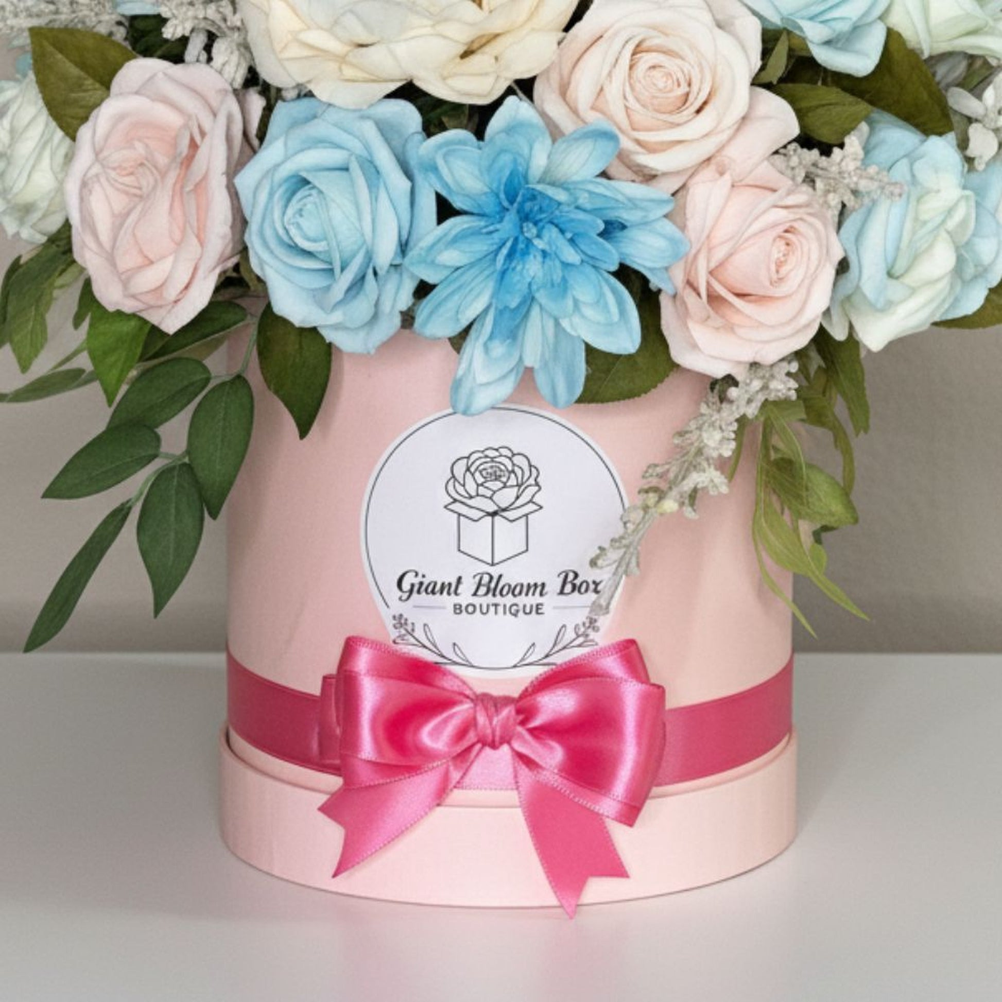 Professional product photograph of an elegant dome-shaped floral arrangement in a light pink cylindrical bloom box. Dense bouquet features artificial roses in soft pastel blue, blush pink, and creamy ivory, along with vibrant sky blue dahlia or chrysanthemum. Dark green eucalyptus leaves on left side, delicate frosted baby's breath filler flowers throughout. The box has wide lustrous hot pink satin ribbon wrapped around it tied in large multi-looped bow at front. White circular label on front displays black