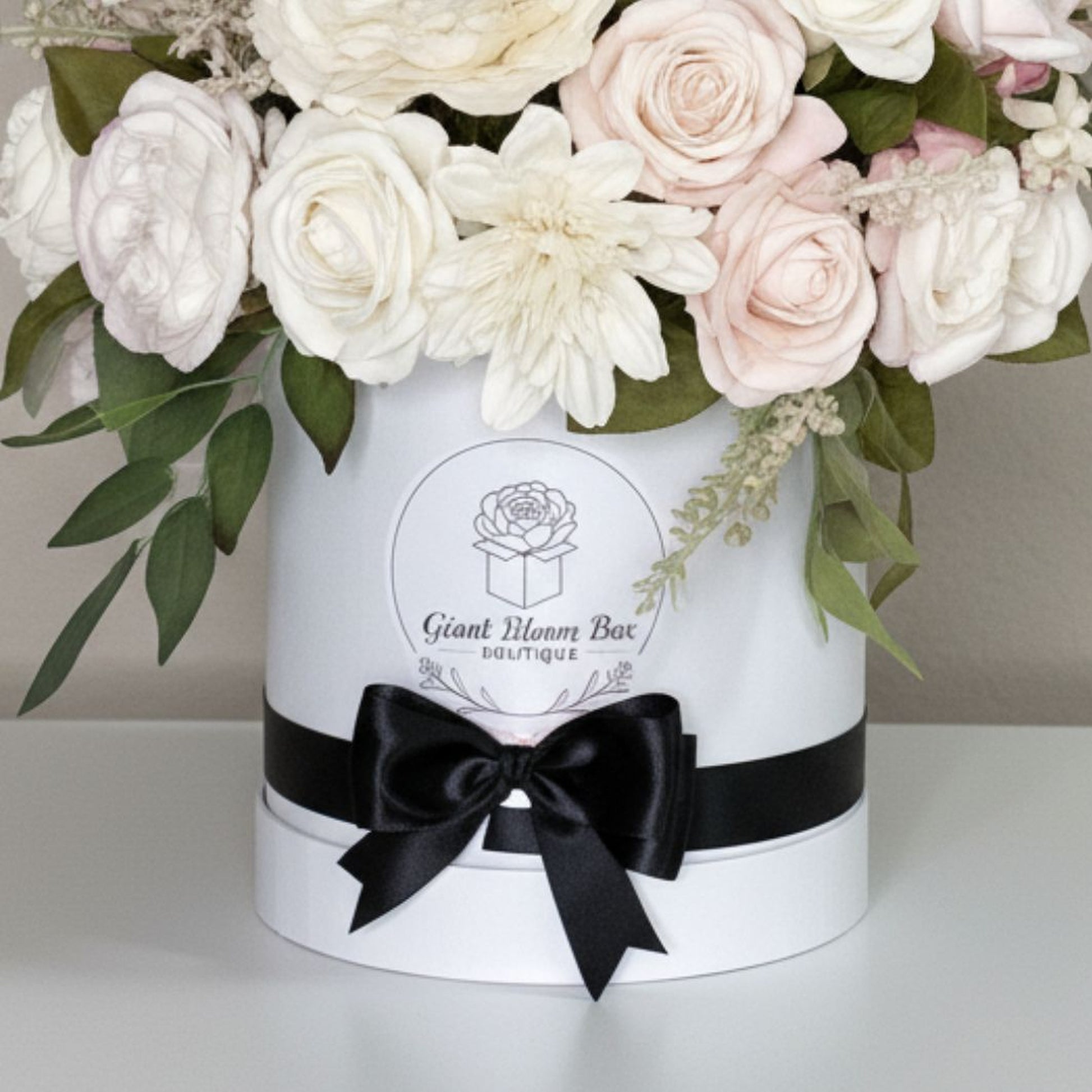 Elegant floral arrangement in round white cylindrical hatbox-style container with separate base and thin rim. Glossy black satin ribbon tied into large bow at front. Centered round logo printed in black line art features stylized rose above open gift box with text 'Giant Bloom Box Boutique' and small decorative flourishes beneath. Arrangement includes many soft pastel blossoms in white, cream, and blush-pink tones: rose-like blooms, prominent large multi-petaled flower resembling chrysanthemum or dahlia in 