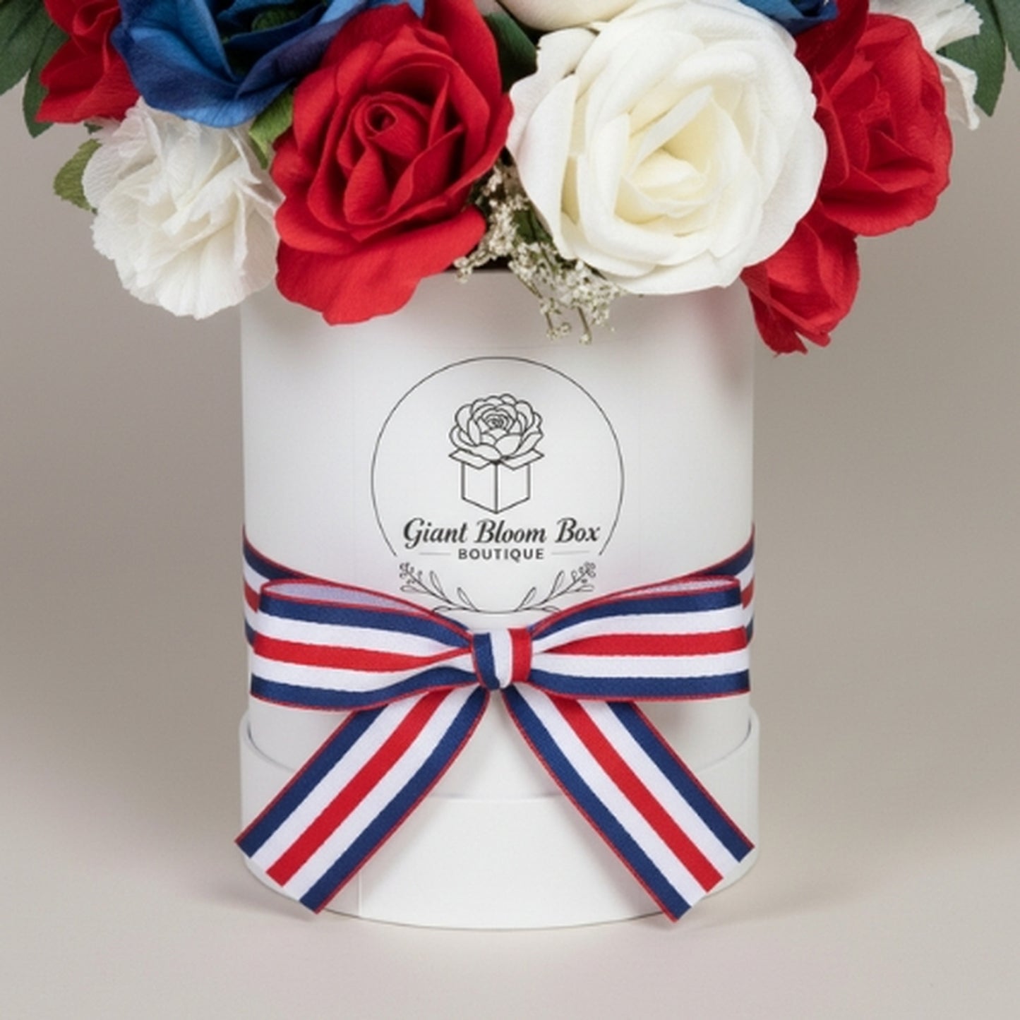 Premium patriotic floral arrangement in white cylindrical Giant Bloom Box boutique container featuring dense dome of red, white, and royal blue artificial roses with baby's breath, branded logo, and striped grosgrain ribbon bow - luxury gift for Memorial Day, Veterans Day, or Independence Day

Keywords: Giant Bloom Box, luxury patriotic flowers, bloom box arrangement, branded flower gift, premium artificial roses, patriotic gift box
