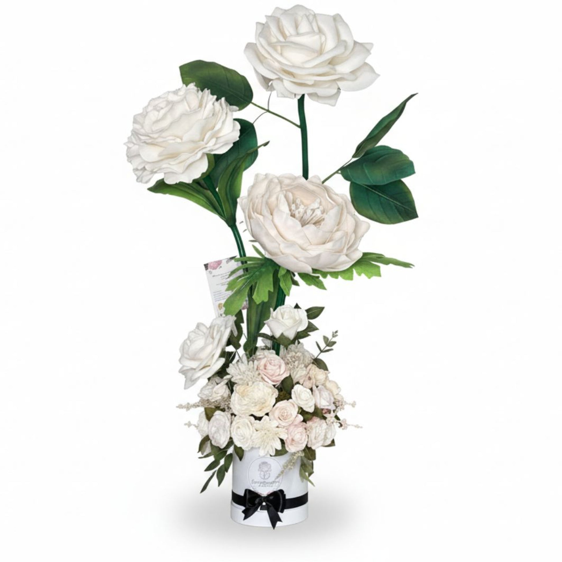 Professional studio product photograph of an elegant tall floral arrangement on pure white background. Three giant oversized white rose-peony flowers rise on long thick green stems with large deep-green leaves at staggered heights above a white cylindrical hatbox container. The base contains a dense bouquet of smaller white and pale blush roses, chrysanthemums, and peony-like blooms mixed with greenery. White container features a printed circular logo, black band near the base, and black satin ribbon tied i