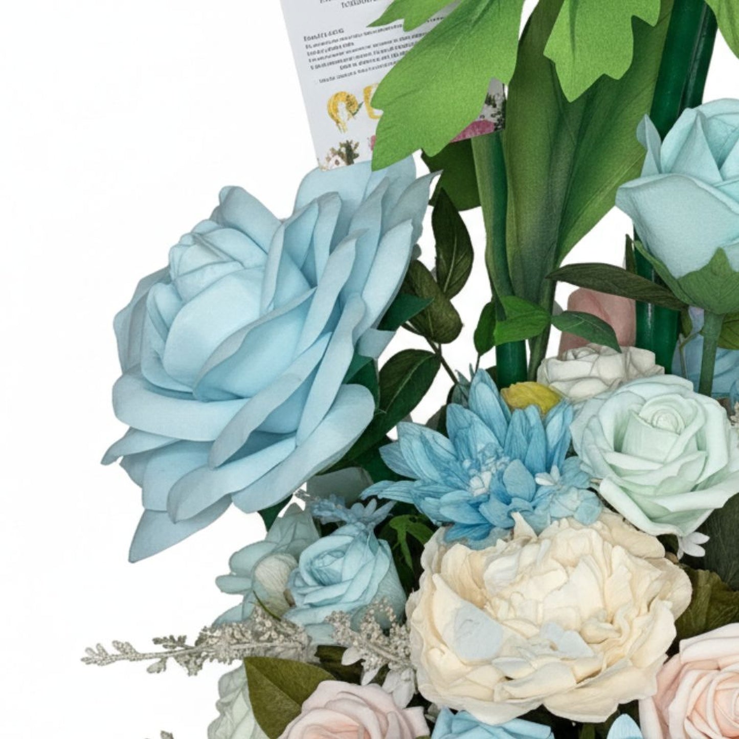 Detailed close-up studio photograph of an artificial paper flower bouquet featuring variety of blooms. Prominent large light blue rose on left side, surrounded by smaller roses in pale blue, mint green, and soft pink. Includes creamy off-white ruffled peony in lower center, vibrant cerulean blue chrysanthemum with spiky petals, and silvery-white baby's breath filler flowers. Dark green leaves with visible veining provide contrast. Small white rectangular card tucked among foliage. White background, bright e
