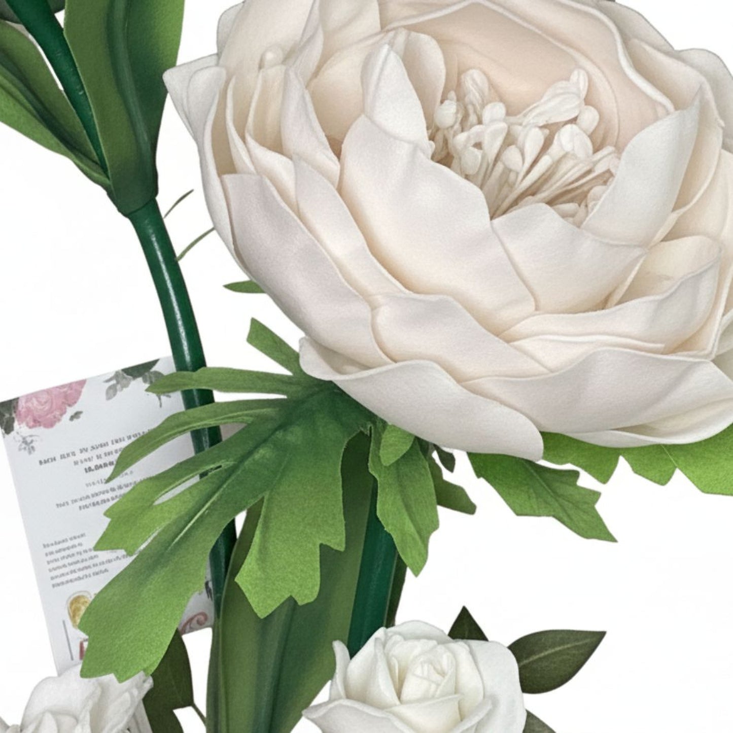 Close-up studio photograph of artificial floral arrangement on clean white background. Large cream-ivory flower resembling peony or fully bloomed rose with many softly curved layered petals and smooth foam-fabric texture. Small white stamen tips clustered in center of bloom. Multiple green leaves in different shapes surround the flower: some broad and smooth, others with deep lobes and serrated cutout edges. Leaves have matte finish, appearing synthetic felt-foam or printed fabric. Thick dark green plastic-