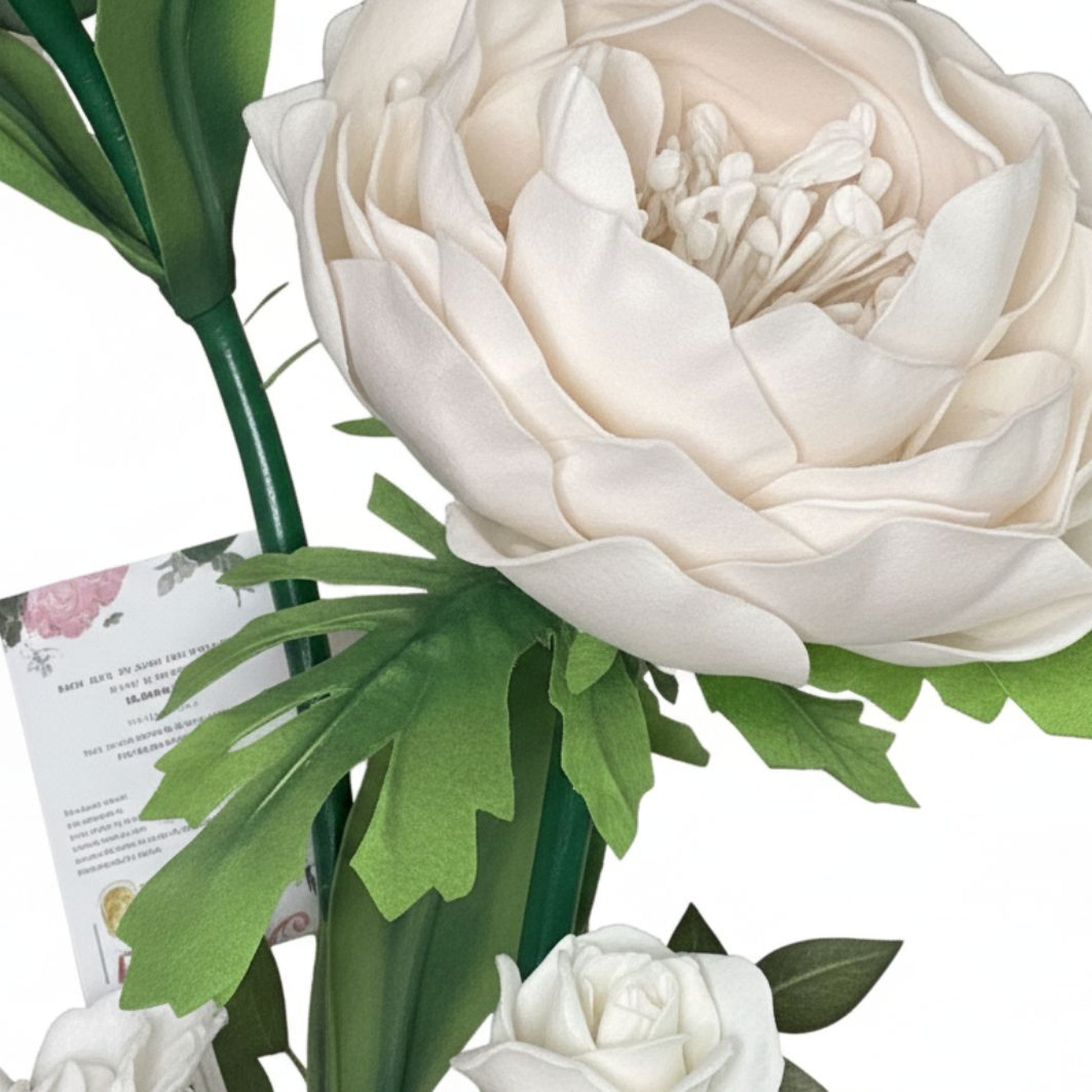 Close-up studio photograph of artificial floral arrangement on clean white background. Large cream-ivory flower resembling peony or fully bloomed rose with many softly curved layered petals and smooth foam-fabric texture. Small white stamen tips clustered in center of bloom. Multiple green leaves in different shapes surround the flower: some broad and smooth, others with deep lobes and serrated cutout edges. Leaves have matte finish, appearing synthetic felt-foam or printed fabric. Thick dark green plastic-