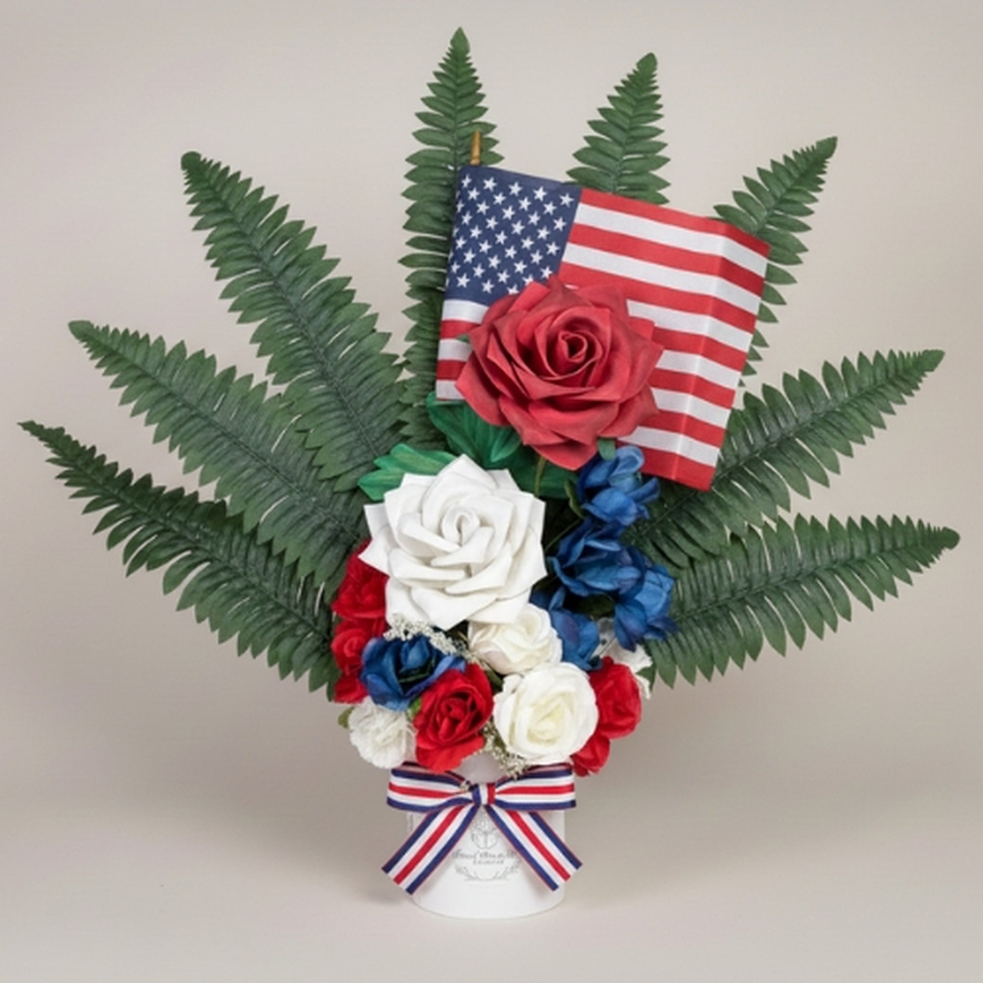 Patriotic artificial floral arrangement featuring red, white, and blue roses with dark green fern backdrop, small American flag, and striped ribbon in white cylindrical vase on neutral background - perfect for Memorial Day, Veterans Day, or Fourth of July commemorationpatriotic flowers, memorial arrangement, American flag decor, red white blue roses, Veterans Day flowers, Memorial Day centerpiece