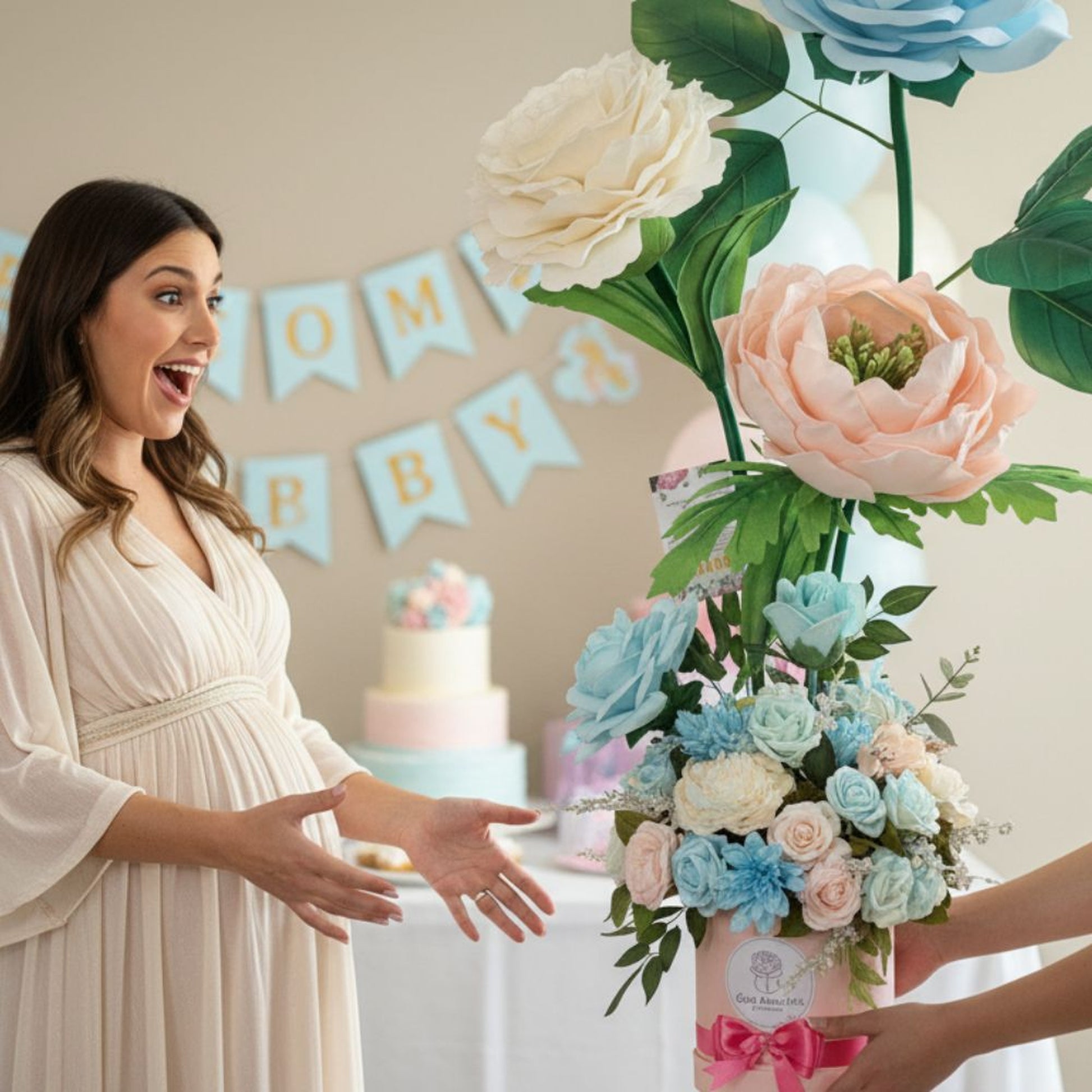 Candid photograph of a pregnant woman in a cream-colored maternity dress expressing joyful surprise with wide eyes and open mouth as she receives a giant floral arrangement at her baby shower. The arrangement features three oversized flowers (light blue rose, creamy white peony, and soft pink peony with green center) in a pink hatbox with bright pink ribbon. Baby shower decorations visible in background include blue pennant banner spelling 'OH BABY', two-tiered pink and blue cake on white tablecloth, and pa