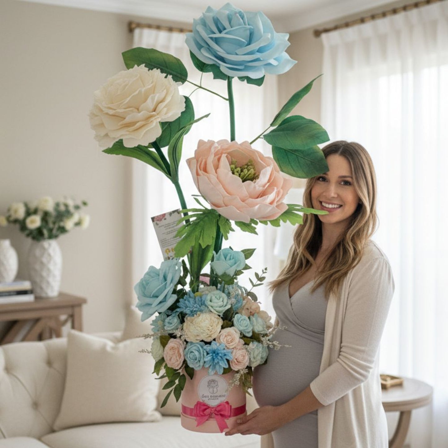Portrait of a smiling pregnant woman with long wavy brown hair wearing a light gray V-neck maternity dress and cream cardigan. She holds an enormous floral arrangement featuring three giant flowers (light blue rose, cream peony, and peachy-pink peony) in a pink hatbox with dark pink ribbon bow. Indoor living room setting with neutral decor, white tufted sofa, wooden console table with white vases, sheer curtains, and soft natural lighting. Professional lifestyle photography showcasing maternity and celebrat