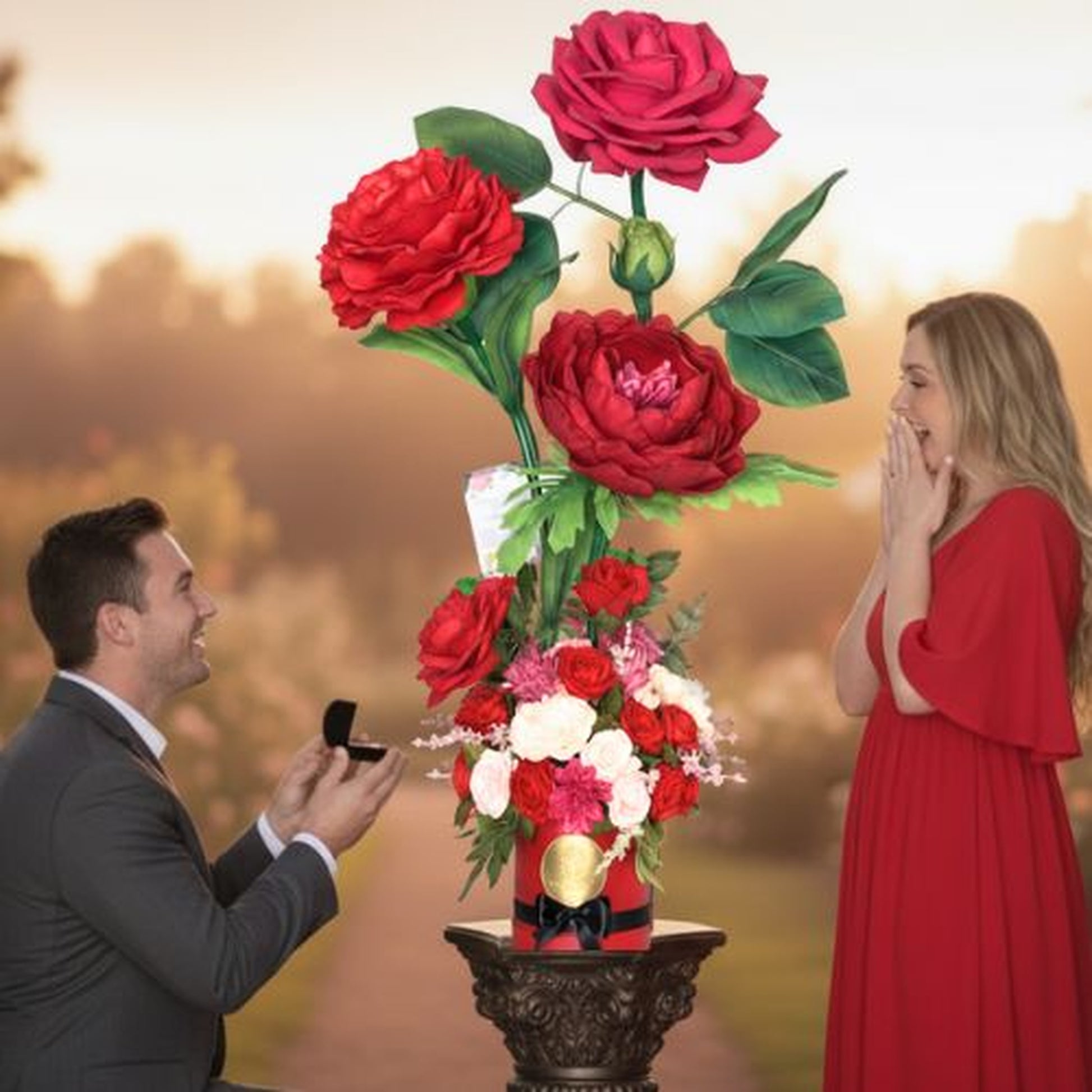 "Romantic outdoor proposal scene captured during golden hour. Man in dark grey suit kneeling on one knee presenting a black ring box to a blonde woman in a flowing red dress. The woman has both hands raised to her face in delighted surprise. Between them is an elaborate floral arrangement on an ornate pedestal featuring three giant red roses in a red hatbox with black ribbon and gold seal. Warm sunset lighting illuminates the park setting with soft-focus trees in the background."