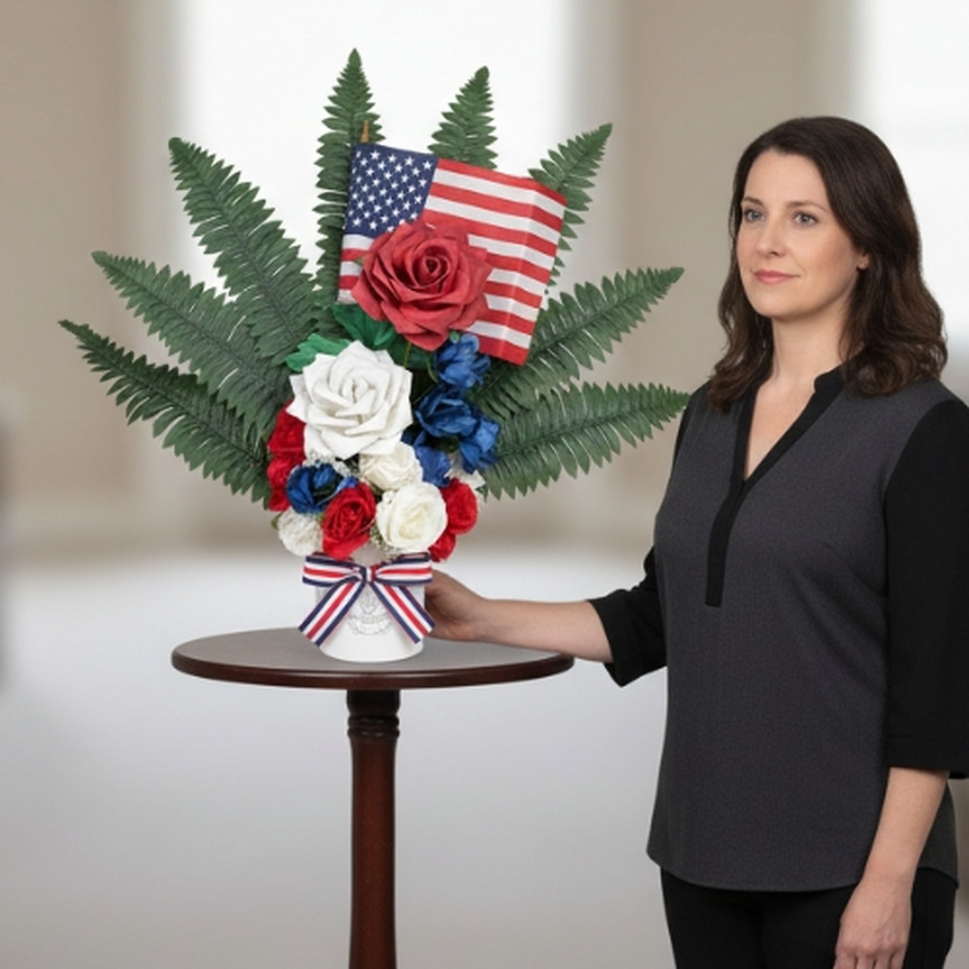 Professional lifestyle photograph showing woman in elegant charcoal gray attire standing beside patriotic floral arrangement on dark wood pedestal table, featuring red, white, and blue artificial roses with American flag and fern greenery in white vase with striped ribbon - commemorative home decor for patriotic holidays

Keywords: patriotic home decor, memorial flowers lifestyle, Veterans Day arrangement, elegant patriotic display, artificial flower centerpiece